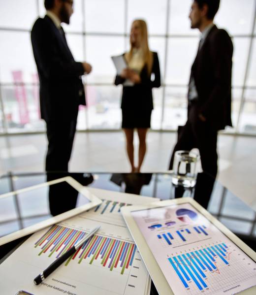 Technological devices and financial document at workplace on background of three business partners discussing ideas